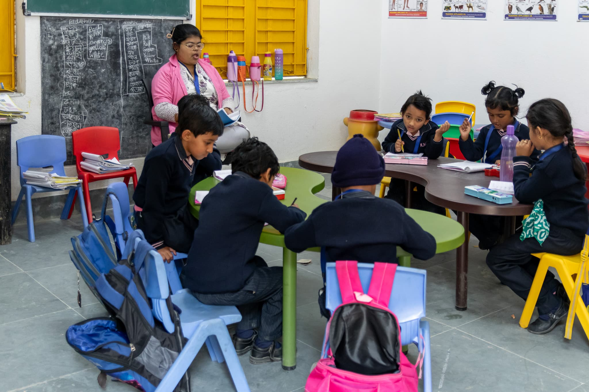 Children working at classroom tables with a teacher nearby